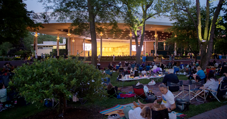 Crowd gathered on lawn with camping chairs at dusk for outdoor concert under illuminated pavilion stage with string lights and tall trees